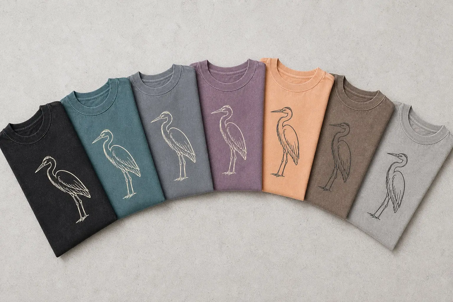 Folded tees fanned in a row across a range of muted grounds, each carrying the same single-colour heron drawing, printed cream on the darker ones and charcoal on the paler ones.