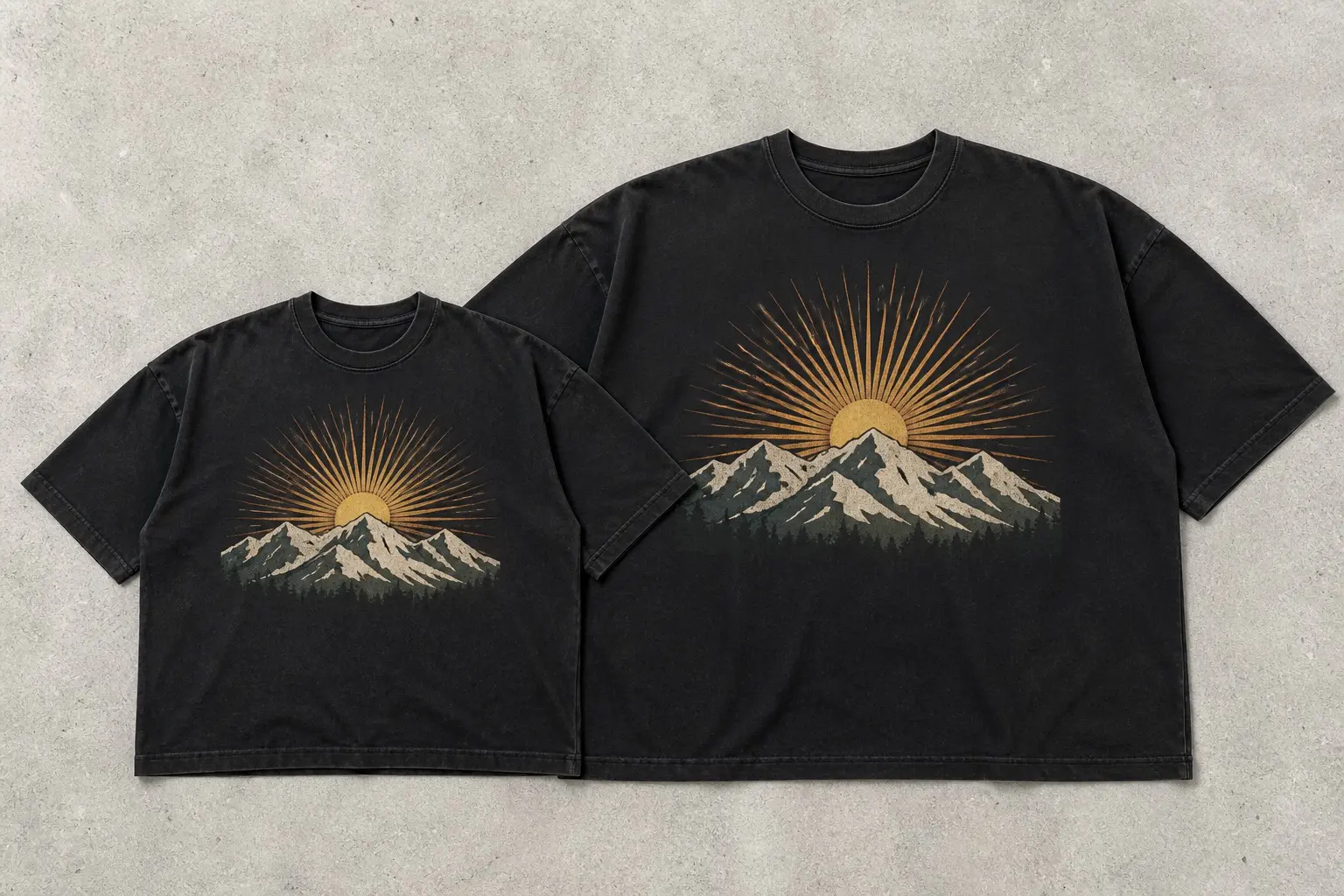 Two black tees laid flat, one small and one much larger, both printed with the same sunburst-and-mountain graphic scaled to cover the same share of the chest.