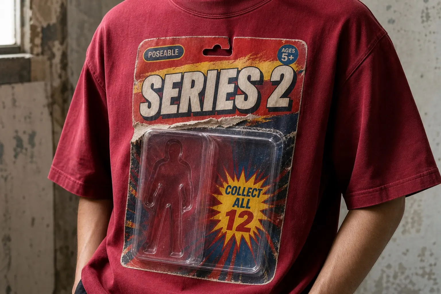A deep red tee worn from the chest down, printed with a toy blister card showing a clear plastic bubble over an empty figure-shaped recess, SERIES 2 across the top, POSEABLE and AGES 5+ in small badges, COLLECT ALL 12 in a yellow starburst, a punched hang hole, and a torn crease down one side.