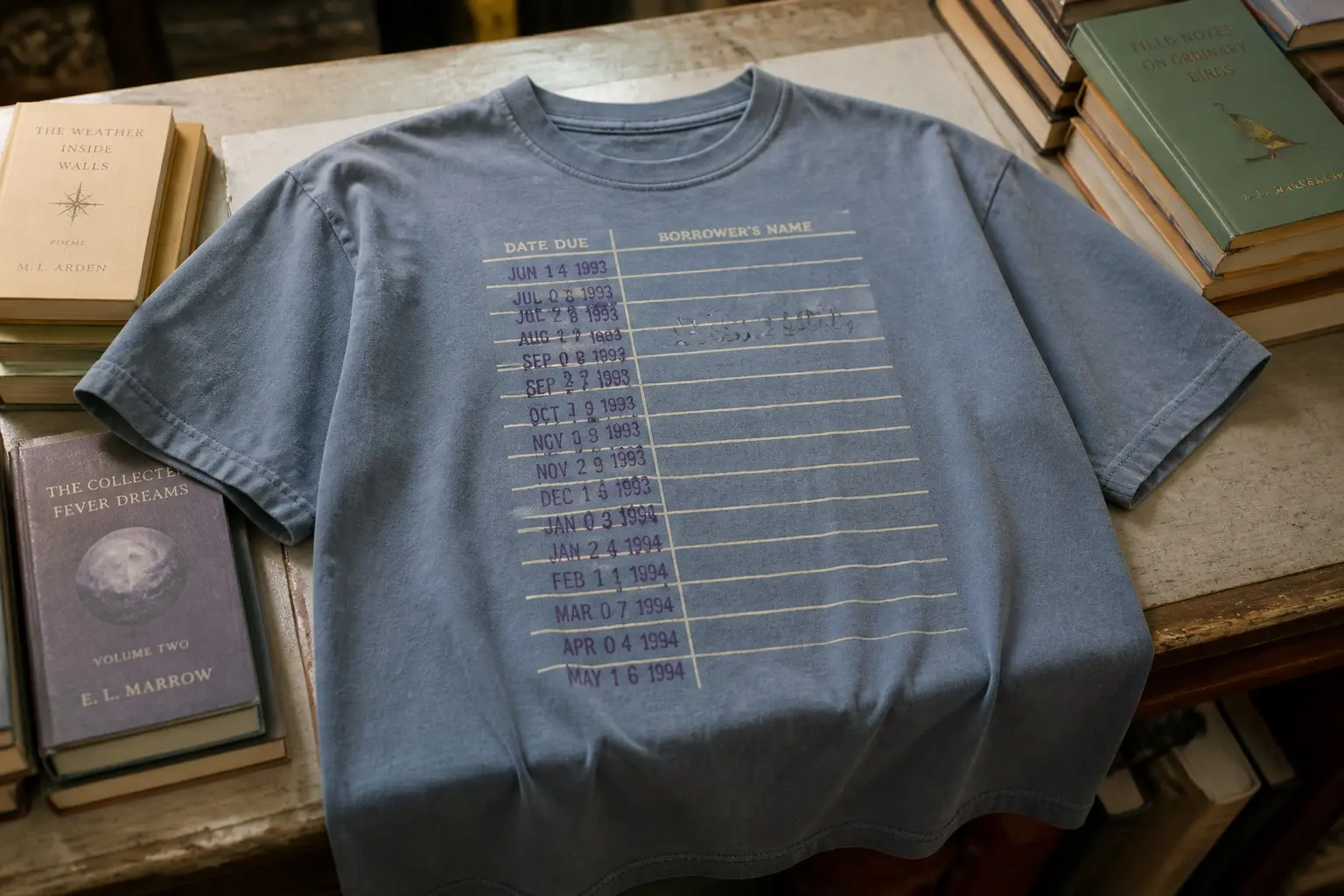 A dust blue tee on a shop counter beside stacked secondhand books, printed with a ruled due-date card carrying a column of overlapping purple date stamps and a handwritten shelf mark