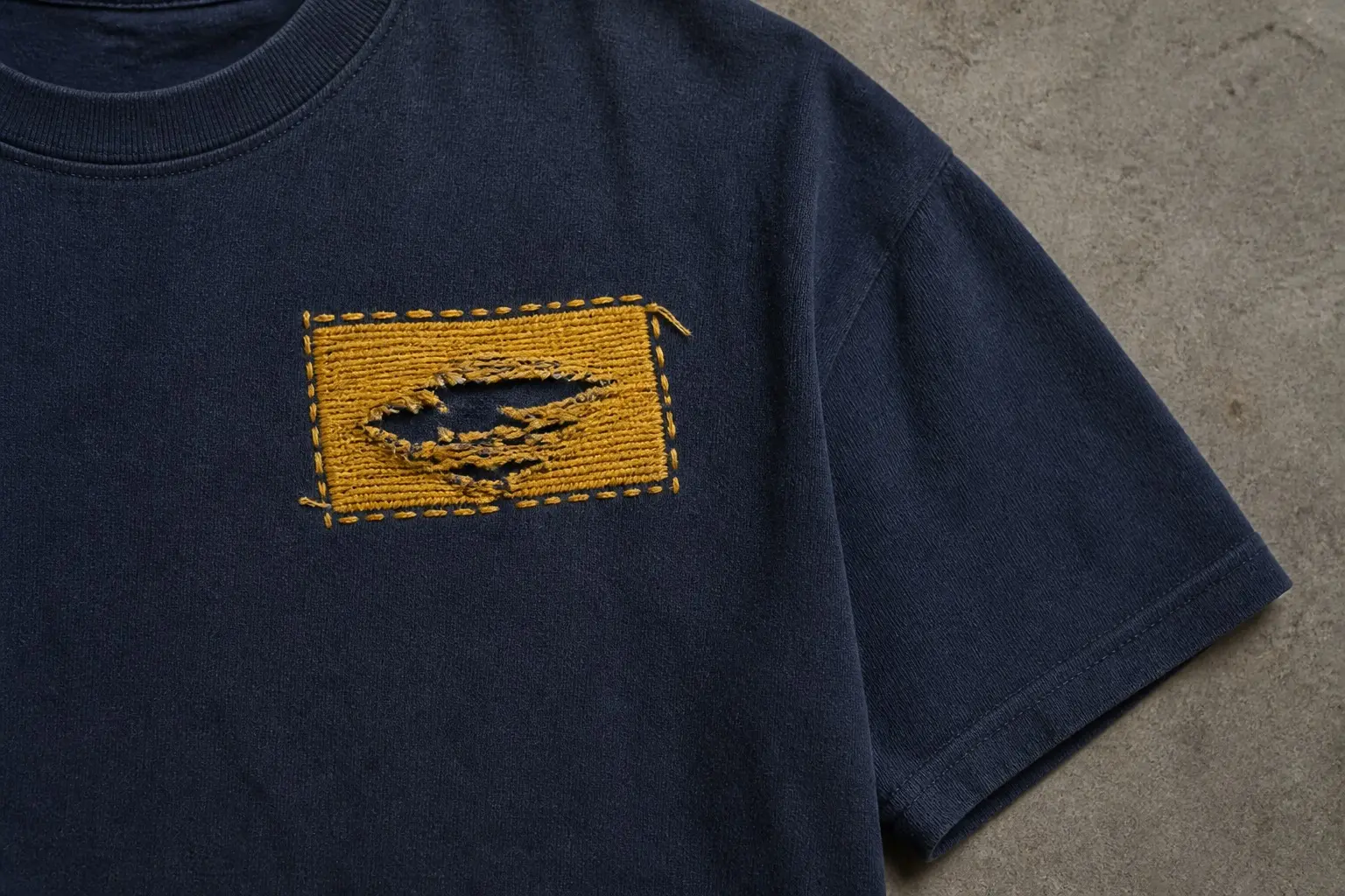 A navy t-shirt shown close on the left chest, printed with a small rectangular mustard patch about the size of a badge, its border an uneven running stitch on all four sides, a frayed slit near the centre and one loose thread left untrimmed at a corner, in a single mustard ink.