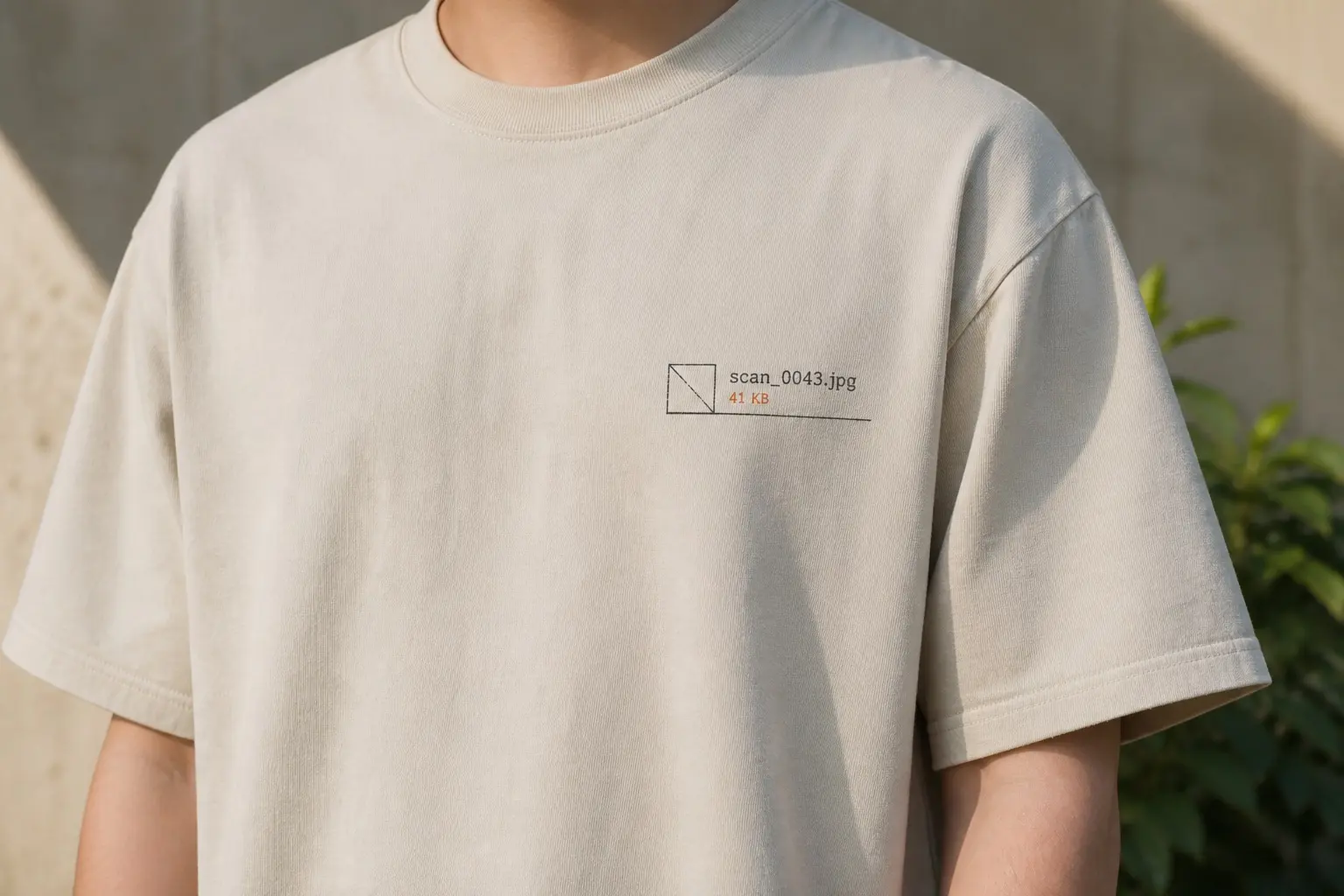 A sand tee with a small badge-sized chest print — an empty outlined thumbnail box crossed by a diagonal line, beside it scan_0043.jpg in dark type over 41 KB in dulled orange, with a hairline rule beneath the whole row.