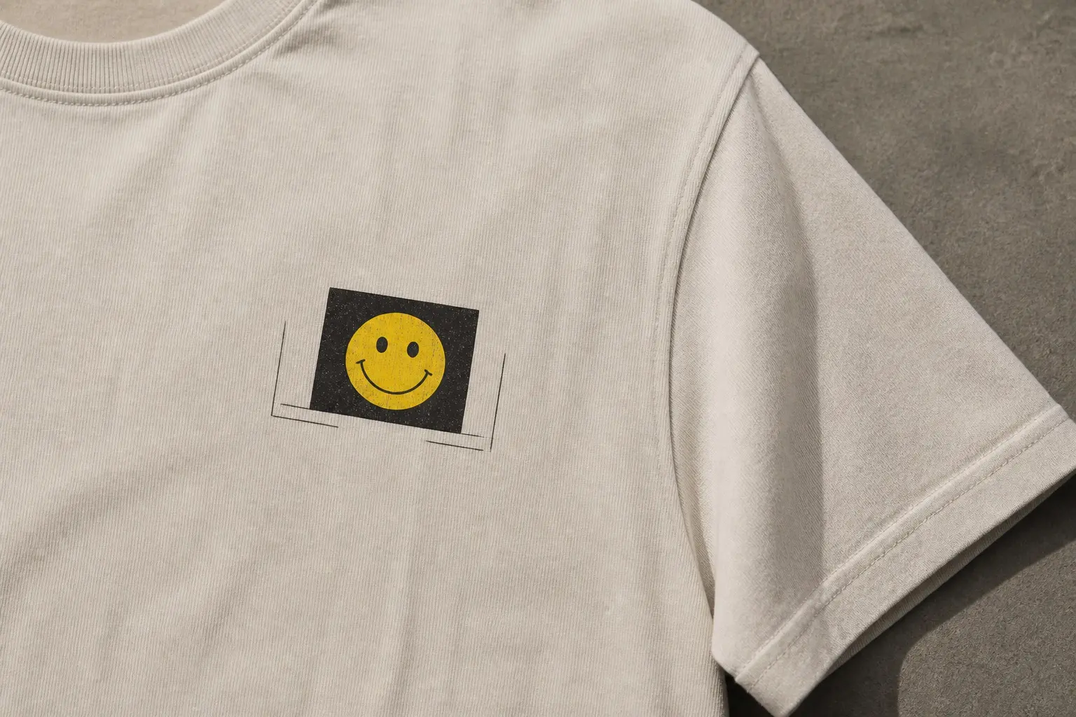 A bone tee laid flat on concrete and shot close on the left chest, printed with a crude yellow smiley inside a hard-edged near-black rectangle tilted a few degrees clockwise, thin dark lines of a room corner running underneath and stopping at the rectangle's edges to pick up again slightly lower on the far side.