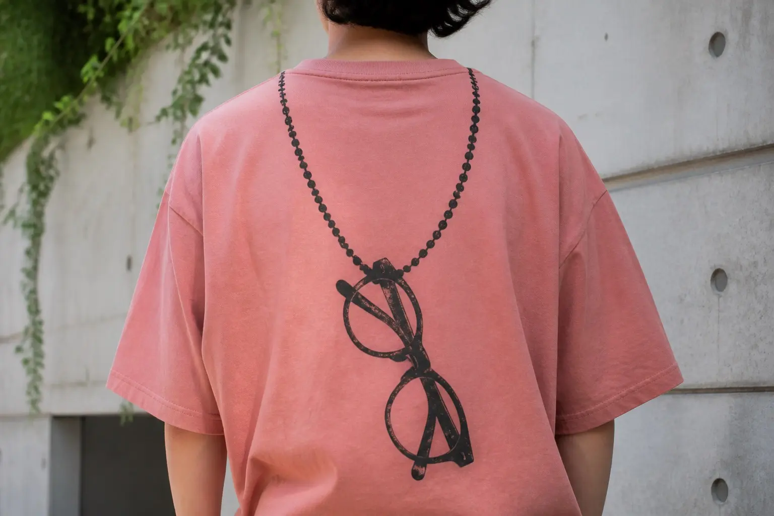A clay pink tee worn from behind and printed across the back, a fine beaded chain running in from the top edge, looping down in a long open curve to a folded pair of reading glasses hanging with one arm crossed over the other, the chain's ends running off the top edge without meeting, no words.