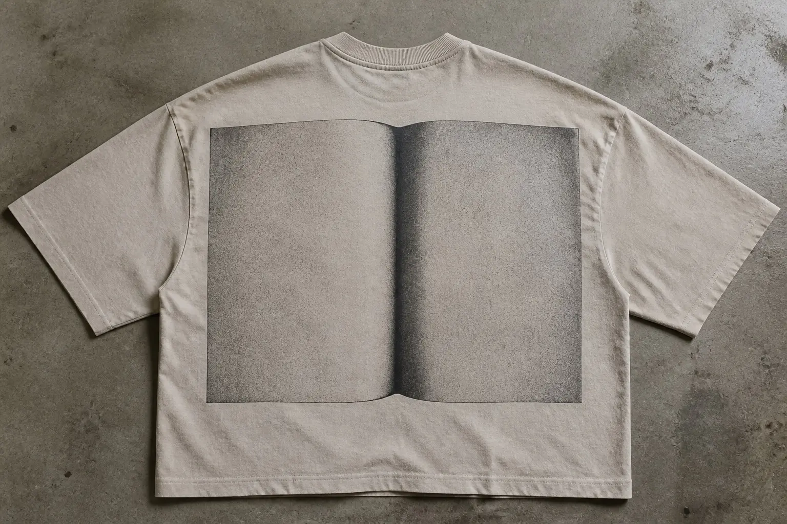 A sand-coloured tee laid flat, back panel printed with a book spread of two blank grey pages divided by a soft dark gradient down the centre where the paper visibly bends away from the binding.