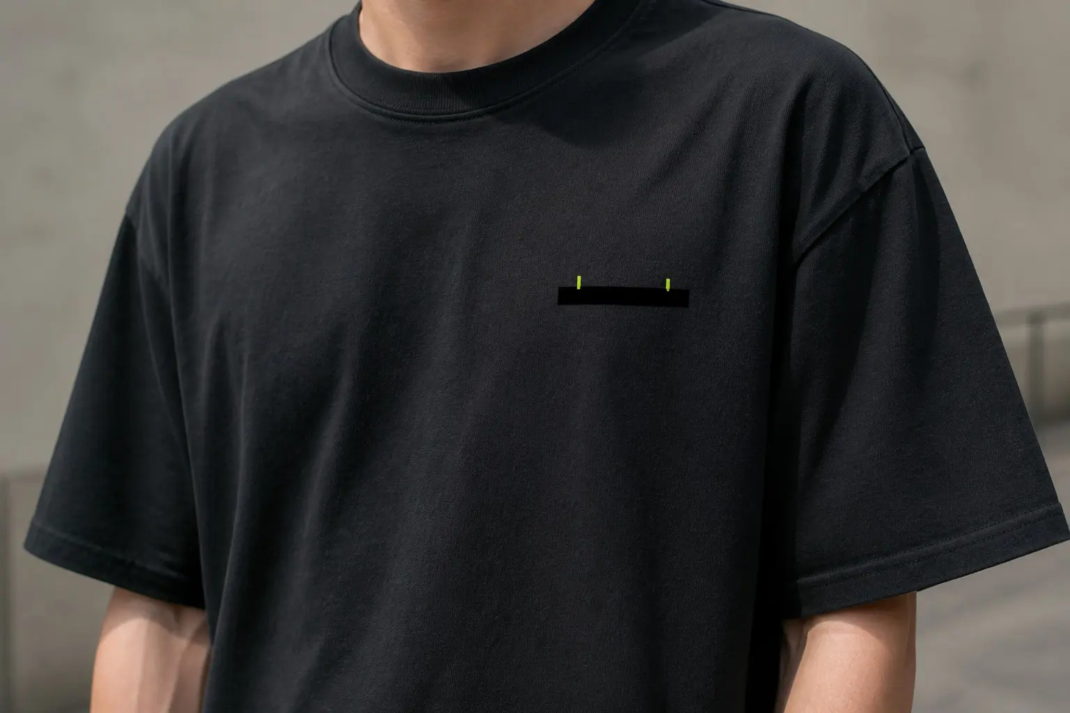 A black tee with a badge-sized chest print — a solid black bar with hard square ends covering one line of text, two lime-green letter tips poking just above its top edge, nothing else in the print.