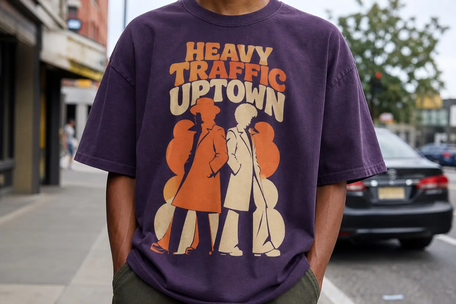 A person from the chest down on a city street, wearing a deep purple tee printed with two full-length silhouette figures in flared coats standing back to back on flat orange and cream blocks, titled HEAVY TRAFFIC UPTOWN