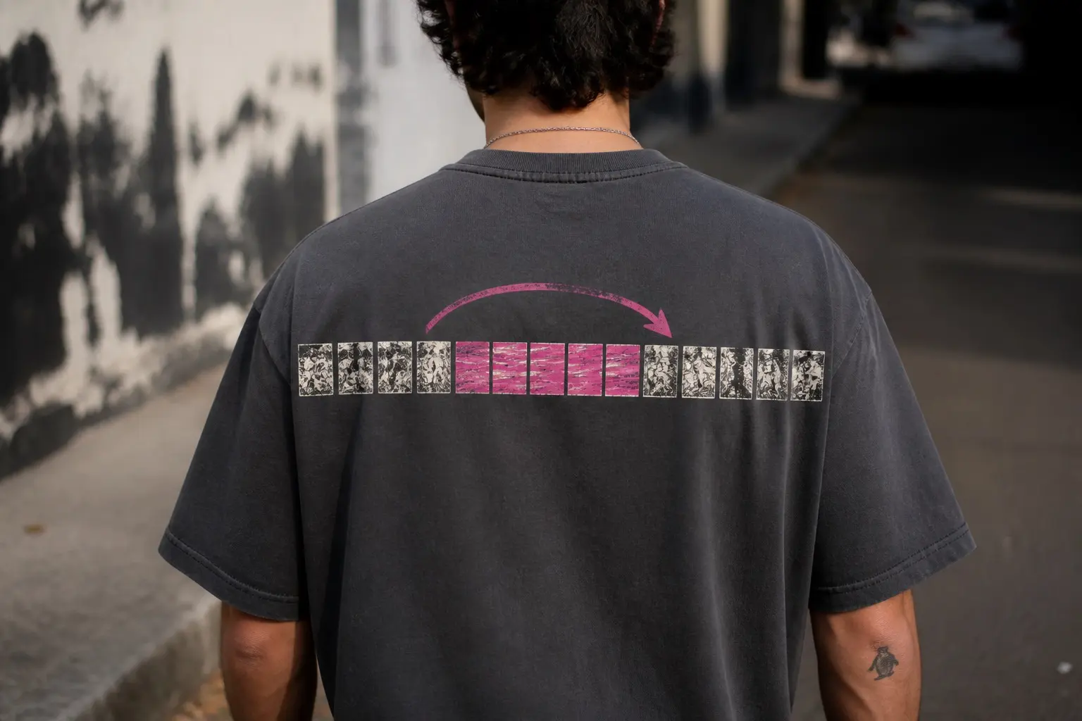 A washed charcoal tee seen from behind on somebody walking away down a sunlit street, printed across the upper back with one unbroken run of fifteen small frames — four of fine black-and-white hatching, then six flat hot-pink frames streaked with hard horizontal lines, then five more hatched — and a bold pink arrow arcing from before the pink frames to just past them.