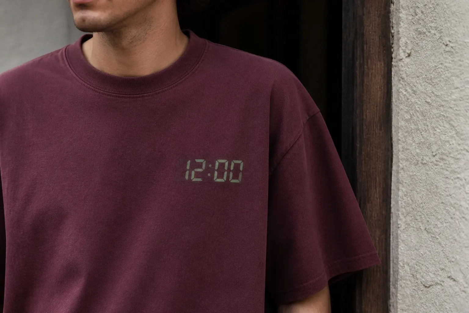 A person standing in a doorway in a deep maroon tee, cropped close on the upper chest, with one small pale green seven-segment display high on the left chest reading 12:00, both dots of the colon lit, a faint halo around the lit segments and the unlit segments showing through as very faint ghost strokes; the rest of the garment is empty.