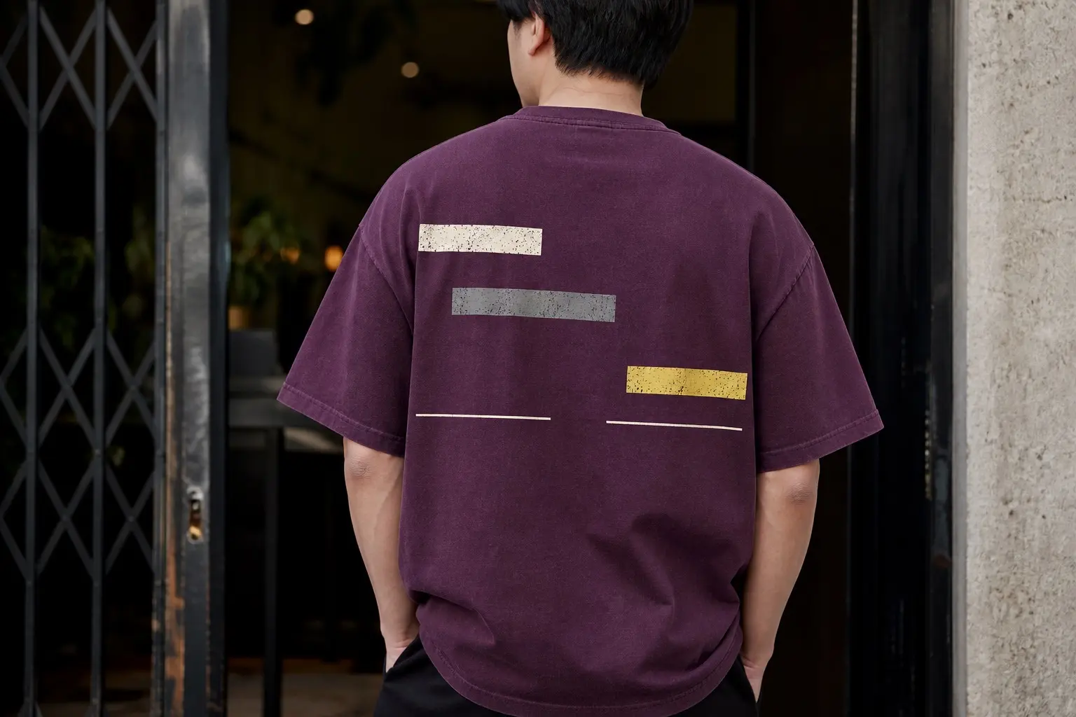A plum tee worn from behind by a person standing in a doorway, printed across the upper back with three short horizontal bars of unequal length — bone white at the top left, mid-grey below it and further right, pale yellow lower and further right again — and beneath them one thin white line running at a constant height in two separate pieces with a clean empty gap between them, nothing at all above that gap. No text.