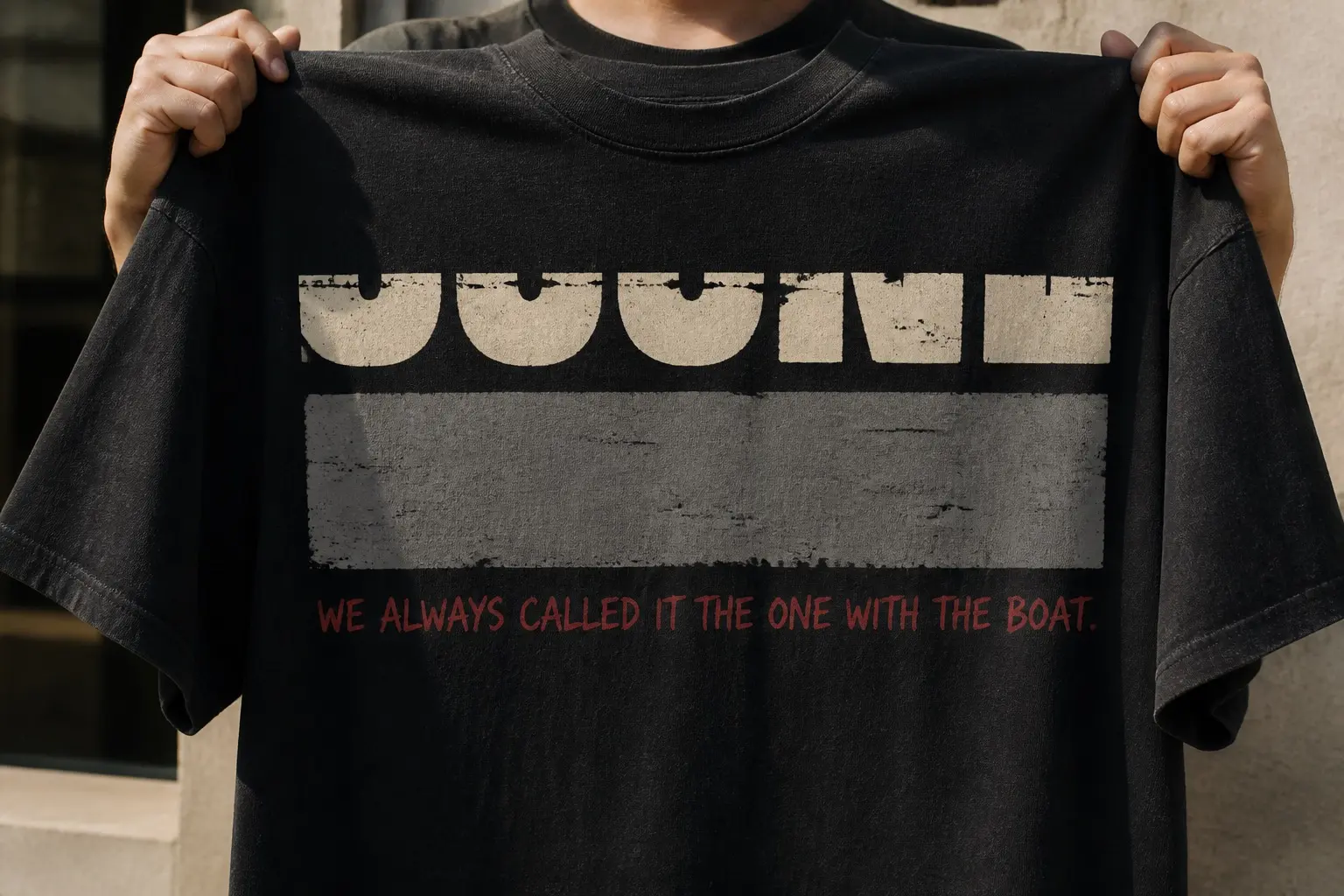 A black tee held up by two hands in front of the wearer, printed with three stacked bands across the chest — cream slab-capital letterforms cropped so only their bottom third survives and nothing can be read, a plain grey rectangle beneath them, and one line of letterspaced dull-red handwriting reading WE ALWAYS CALLED IT THE ONE WITH THE BOAT.