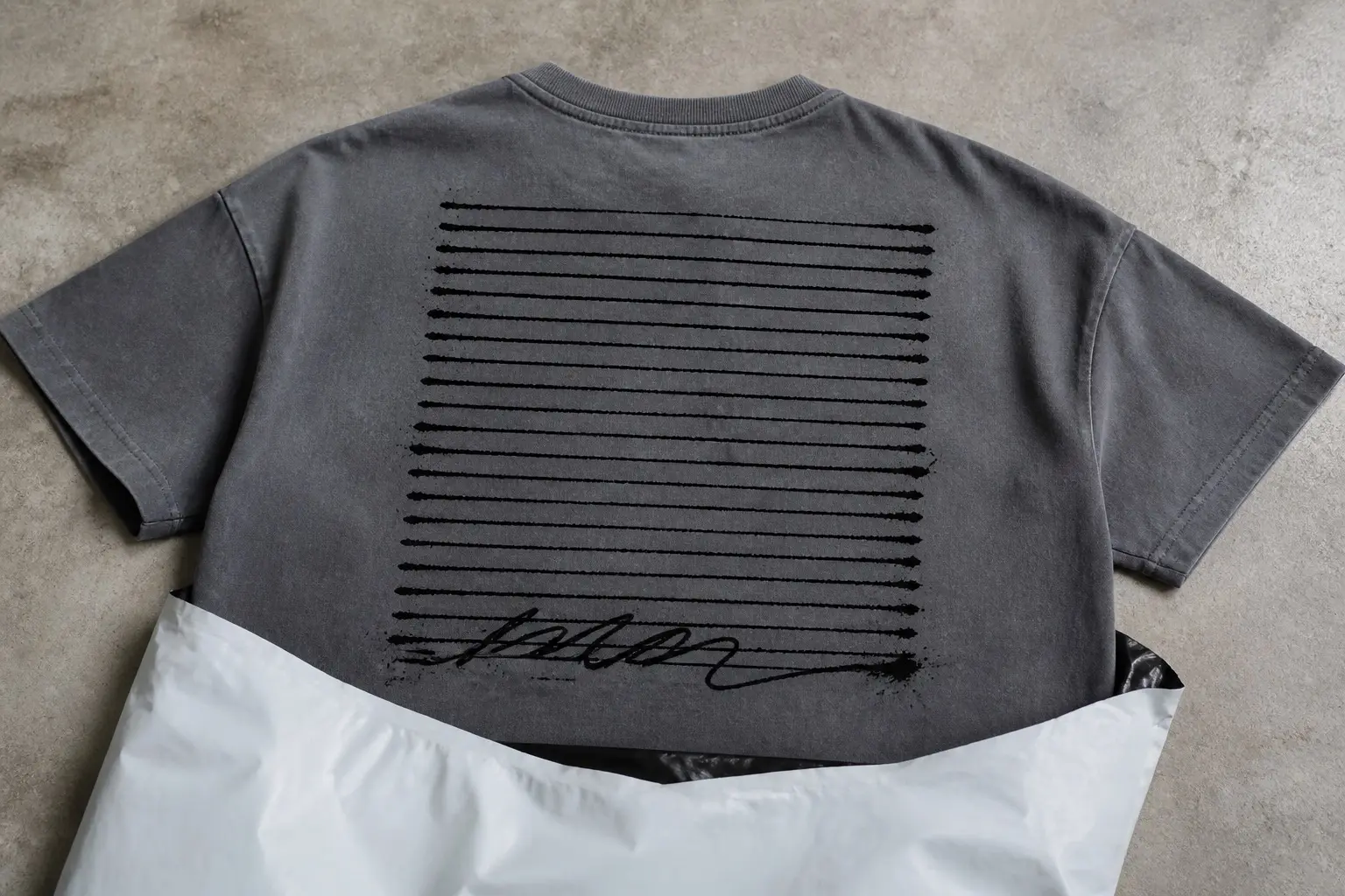 A slate grey tee half out of a shipping bag, printed across the back in black ink with a tall block of blank ruled lines running shoulder to shoulder, every line empty except the bottom one, which carries one wild, illegible looping signature scrawl.