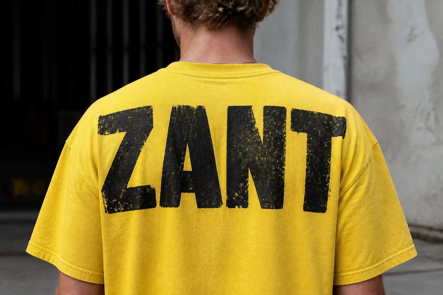 A safety-yellow tee worn from behind, printed with four invented capitals reading ZANT very large across the upper back in heavy plain block form in soot black with a hard keyline — wide letters, open counters, thick even strokes, nothing decorative.