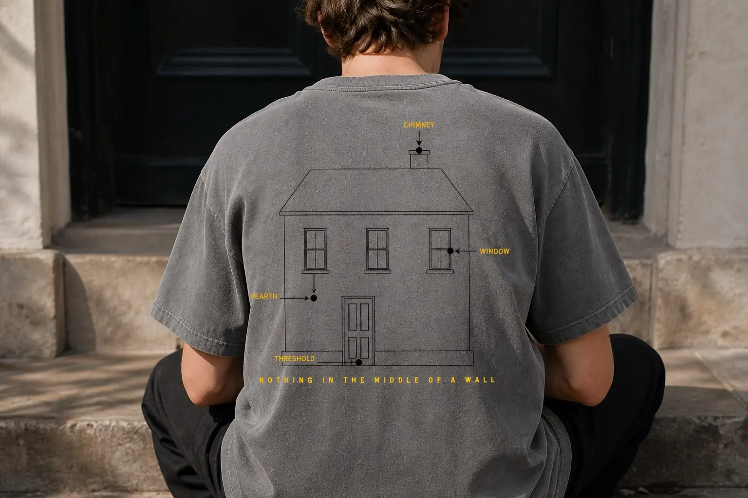 A washed grey tee worn by a man seated on a stone step facing away, back-printed with a thin black ruled elevation of a house — one panelled door, three windows, one chimney, no shading — where four yellow words outside the walls each fire an arrow inward to a small solid black dot at the chimney, the hearth, a window and the door sill, with letterspaced yellow capitals along the bottom reading NOTHING IN THE MIDDLE OF A WALL.