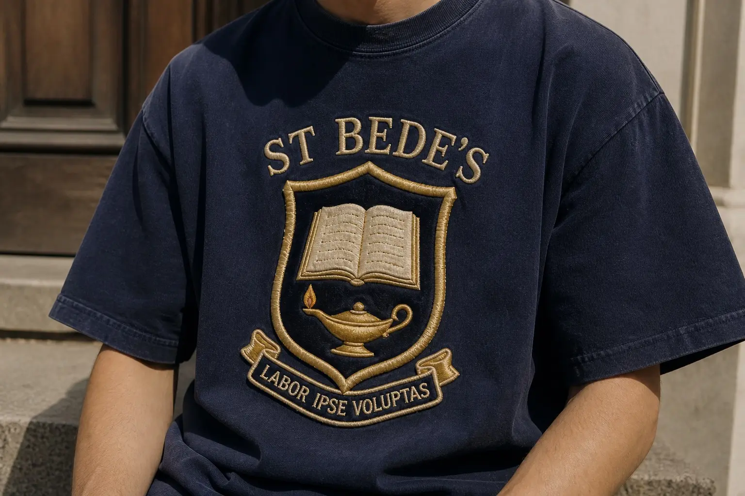 A navy tee worn from the chest down, printed with an embroidered blazer badge in dense satin stitch with a raised bullion border, a plain shield carrying an open book and a lamp, ST BEDE'S arched above and LABOR IPSE VOLUPTAS on a small stitched banner beneath