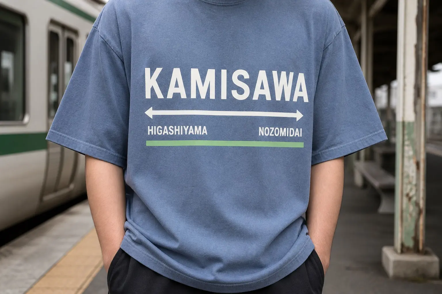 A person from the chest down on a platform, wearing a dust blue tee printed as an enamelled station name board reading KAMISAWA large and centred, with a rule beneath and small arrows to HIGASHIYAMA and NOZOMIDAI