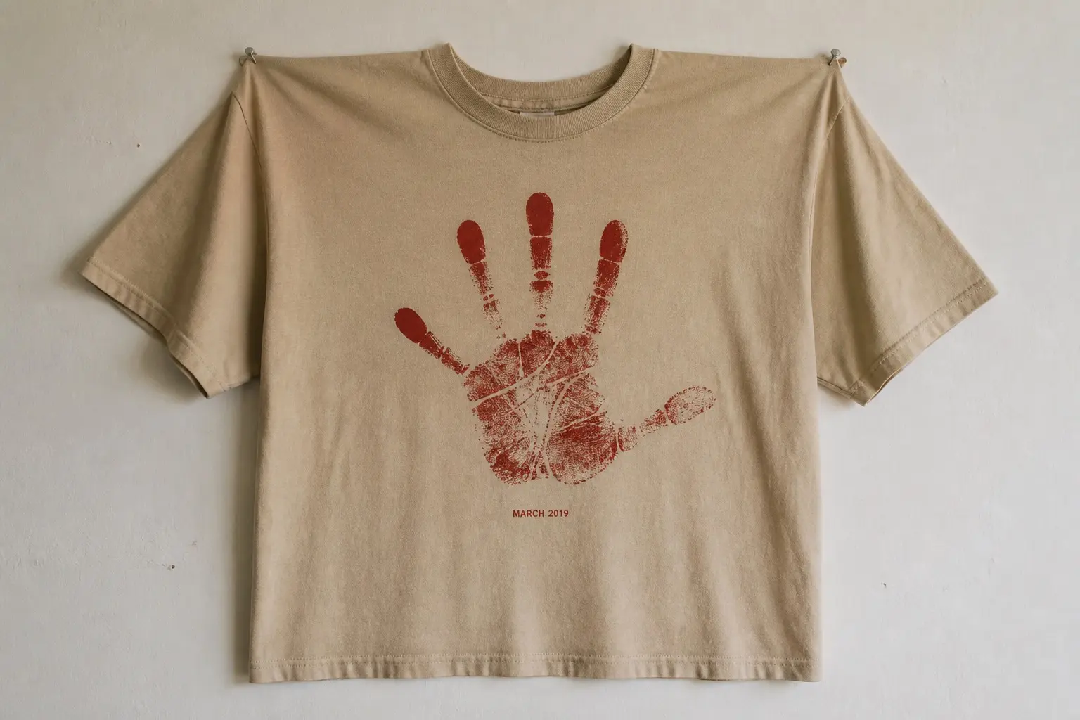 A sand tee pinned flat to a wall by its shoulders, printed with one deep red poster-paint handprint filling most of the chest, fingers splayed, the paint coverage patchy and the palm creases showing as bare lines, MARCH 2019 printed small beneath it.