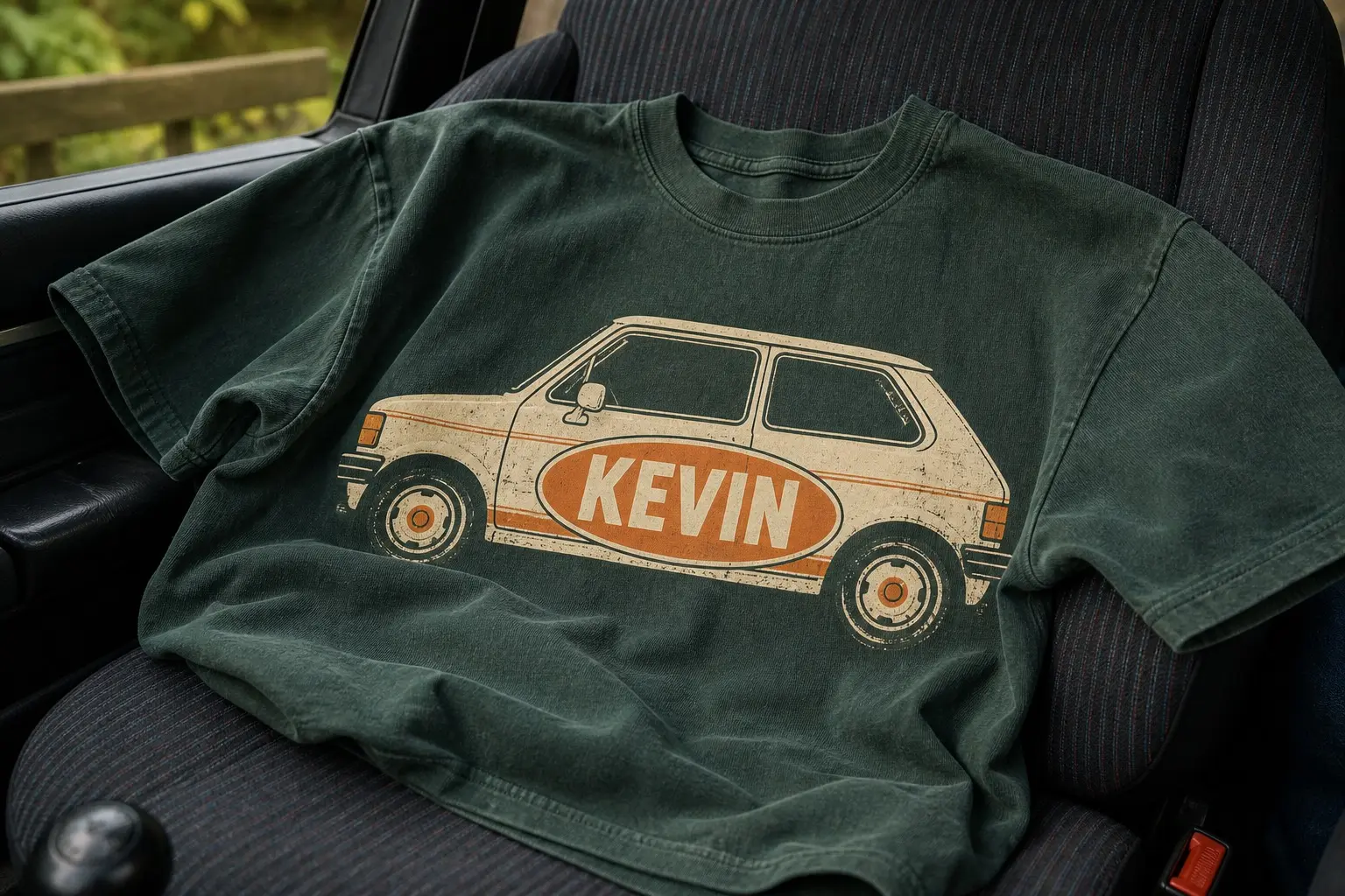 A forest green tee thrown across a car seat, printed with a small cream hatchback in flat side profile and, fixed to its door, an oversized orange oval enamel nameplate reading KEVIN.
