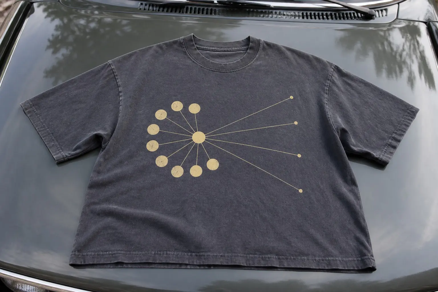 A slate t-shirt spread flat on a car bonnet, printed with a pale yellow hub and thin radiating lines — a tight cluster of eleven large dots close on one side and four small dots a long way off on the other, with no words anywhere.