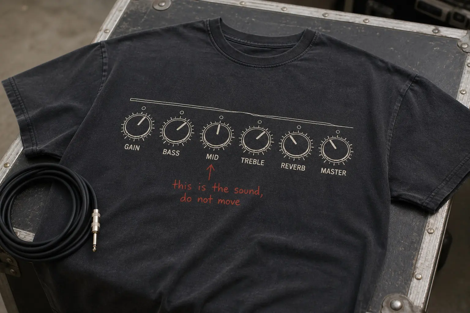 A charcoal tee flat on a flight case beside a cable, printed with a line-drawn amplifier control panel of six labelled knobs each showing a pointer at a specific angle, GAIN BASS MID TREBLE REVERB MASTER printed beneath, and this is the sound, do not move written across the panel in scarlet biro with an arrow to the MID knob.