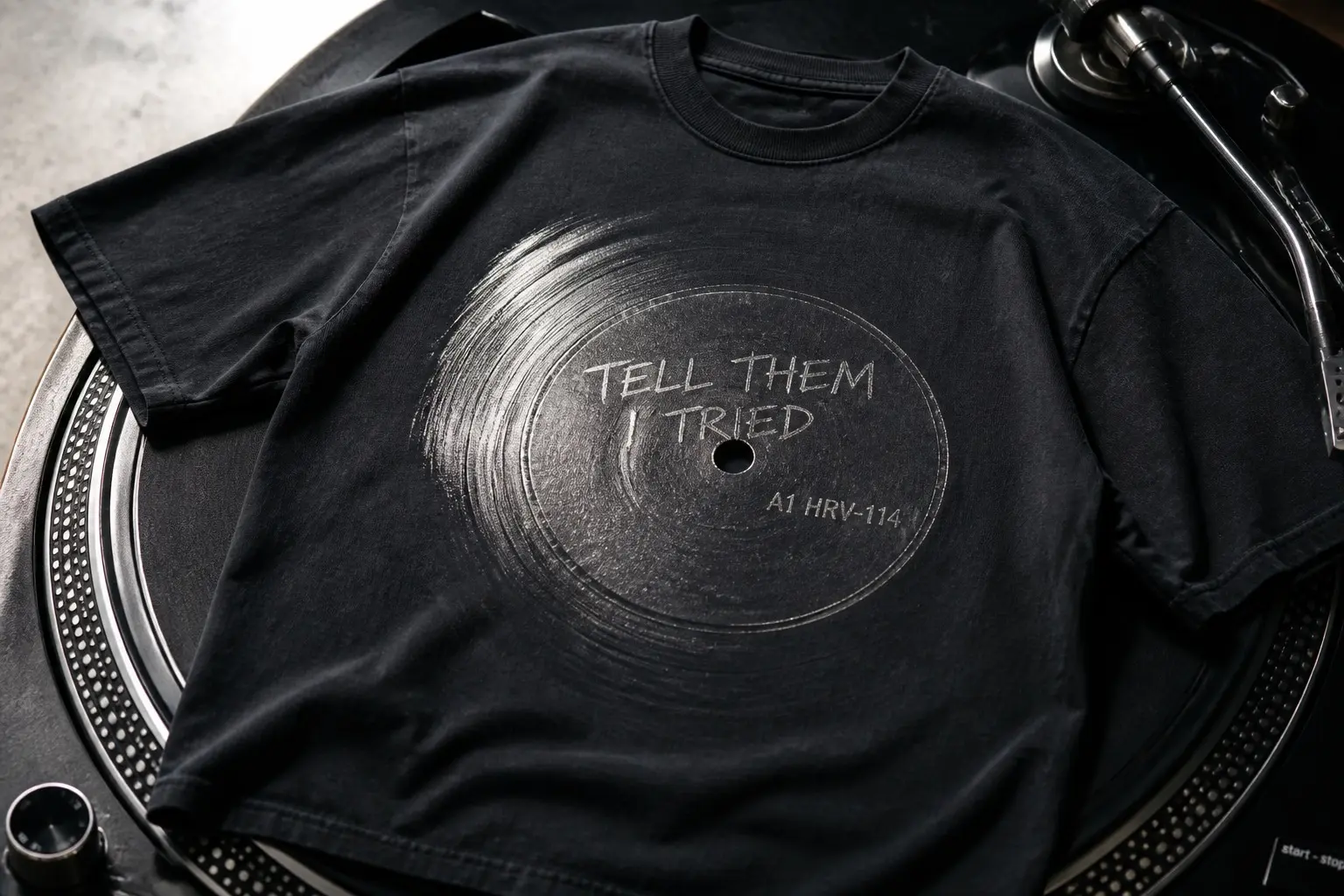 A near-black tee laid flat on a turntable platter, printed with the blank run-out area of a record showing TELL THEM I TRIED scratched into it in uneven hand cursive, the matrix code A1 HRV-114 stamped beside it, and groove lines curving away