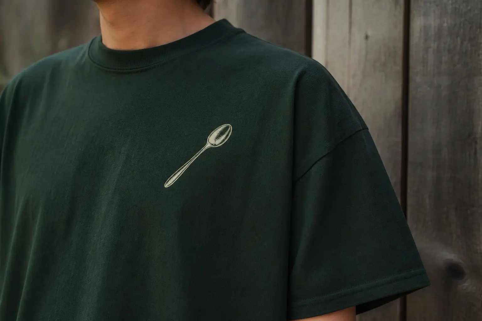 A forest green tee worn outdoors near a wooden fence, printed on the chest with a single kitchen spoon in fine cream engraved line, small and set toward the centre, the rest of the fabric bare.