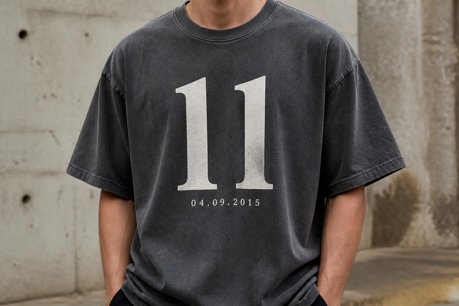 A charcoal tee worn from the chest down, printed with the numeral 11 set very large and plain in the centre of the chest and the date 04.09.2015 in small letterspaced type well beneath it, with nothing else on the garment