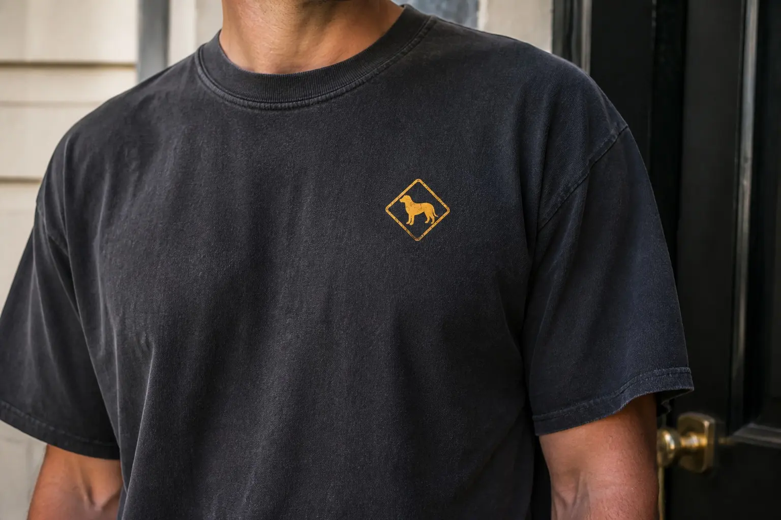 A charcoal tee worn near a dark front door with a brass handle, printed with one small distressed amber diamond high on the left chest holding a plain dog silhouette standing square in profile, ears relaxed, tail level, with no wording anywhere on it.