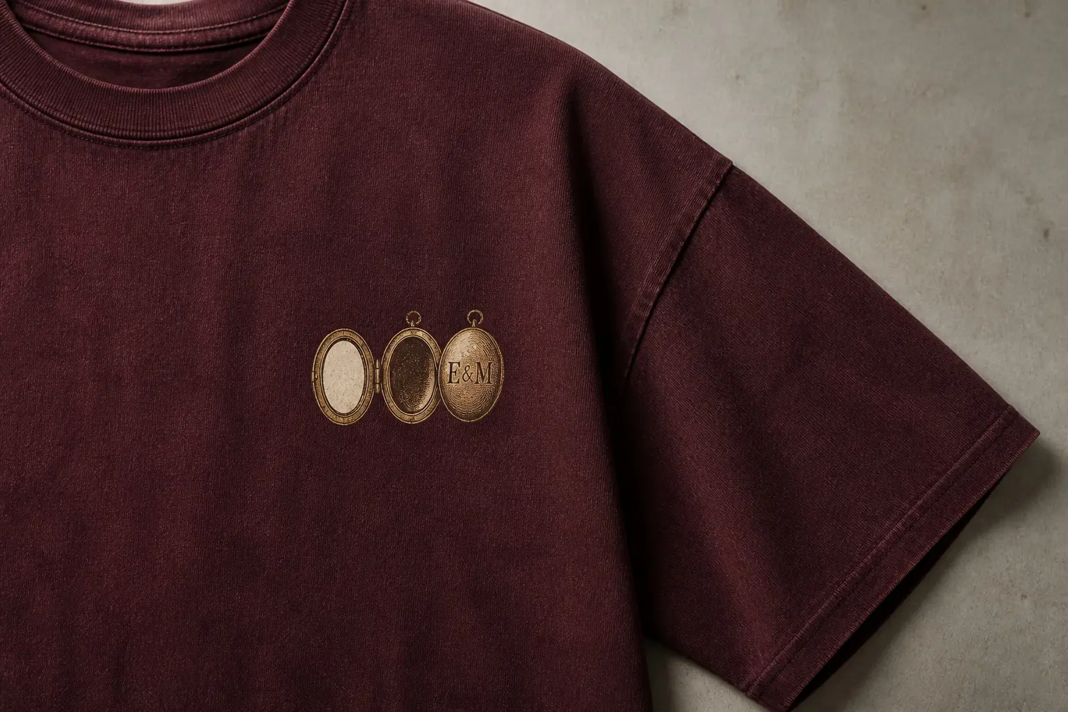 An oxblood t-shirt printed with a small left-chest mark in bone and antique brass ink, an oval locket open on its hinge with one interior face holding a small blank oval cut to fit and the other interior face's backing left bare, and beside it the same locket's case drawn closed and engraved E & M.