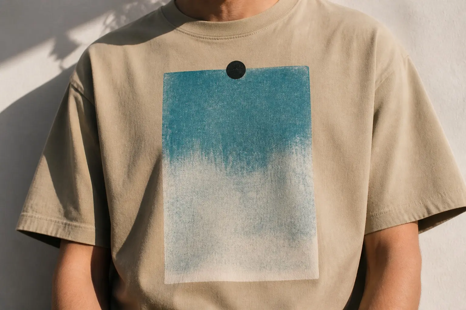 A warm sand t-shirt worn from the chest down, printed large and centred on the chest with a single blank photographic rectangle held at its top edge by one round black magnet, washed teal-blue at the top and fading unevenly to near-white toward the bottom.
