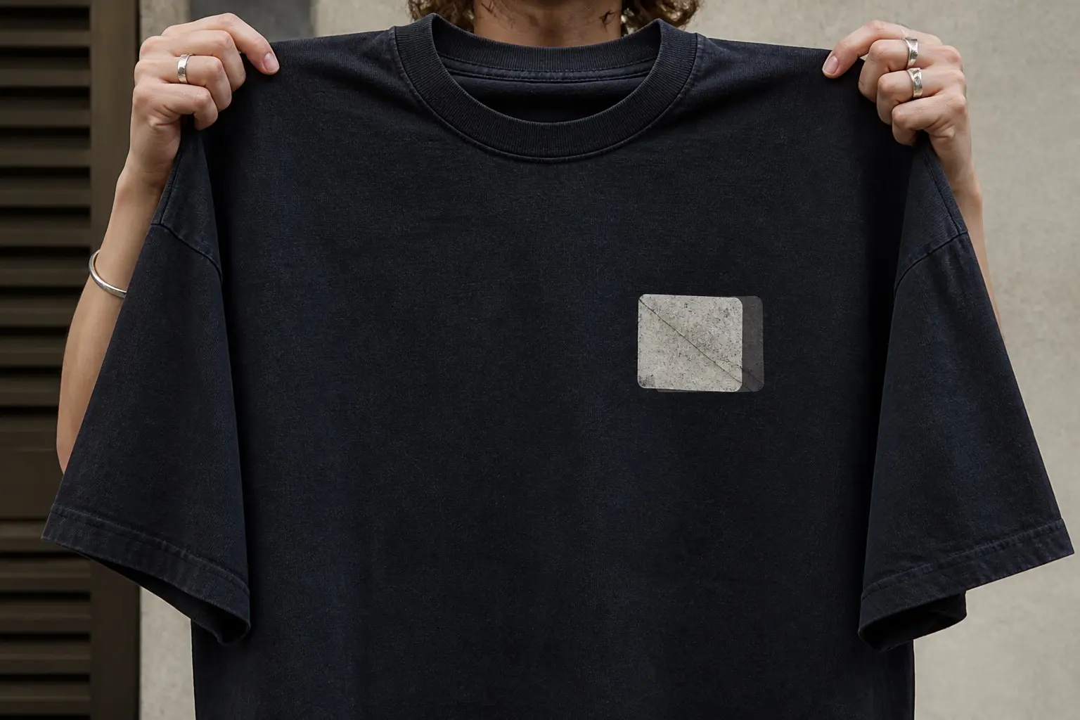 A near-black t-shirt held up by two hands in front of the person wearing it, printed high on the left chest with one small blank photograph roughly nine centimetres across, its corners rounded soft, one diagonal crease running corner to corner, sitting half withdrawn from a plastic wallet sleeve whose edge trails behind it.
