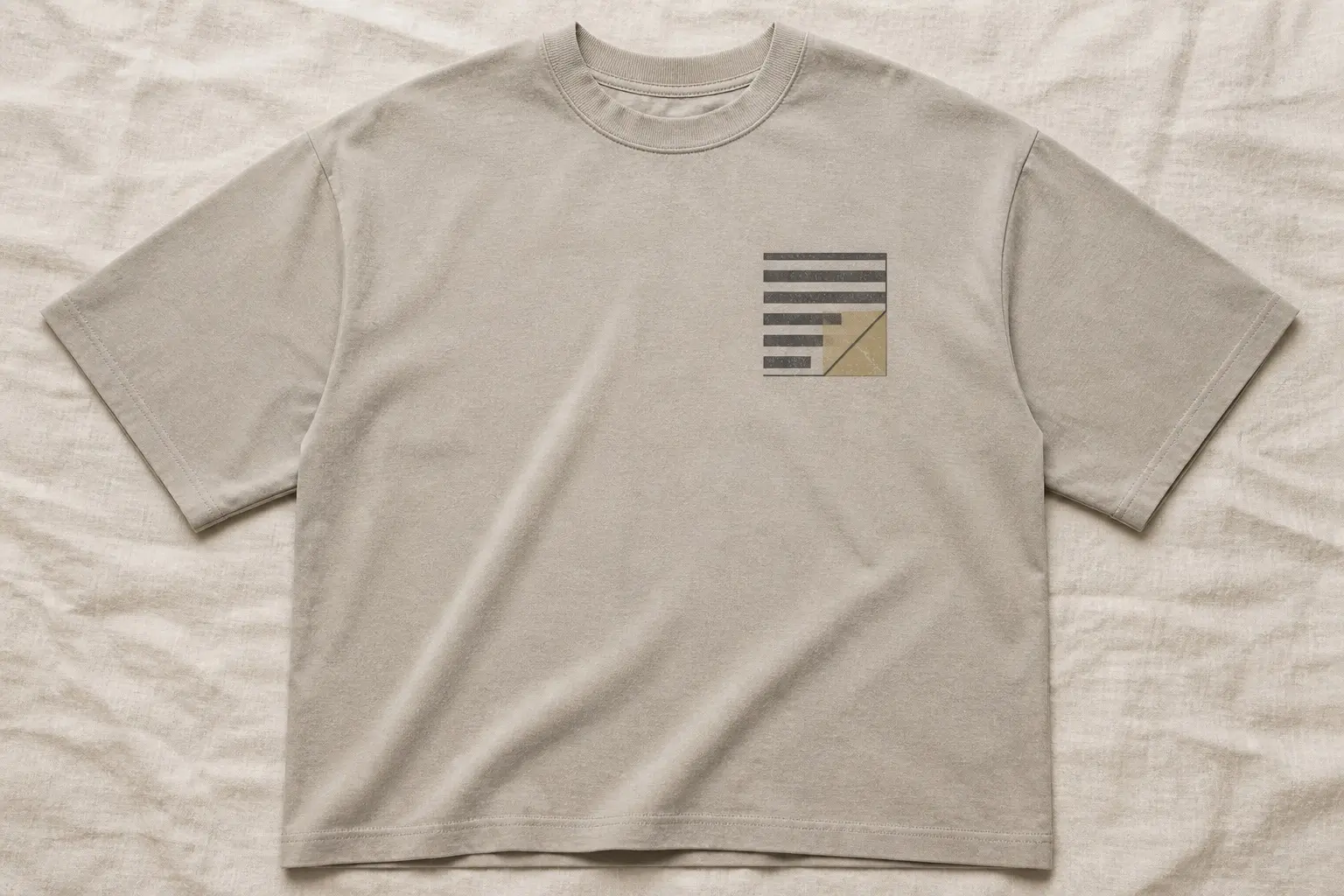 A stone-coloured tee shot flat on a linen sheet, printed small and high on the left chest with a square page corner — grey bars of type across the upper left, a tan folded region in the lower right, and one hard diagonal crease between them.