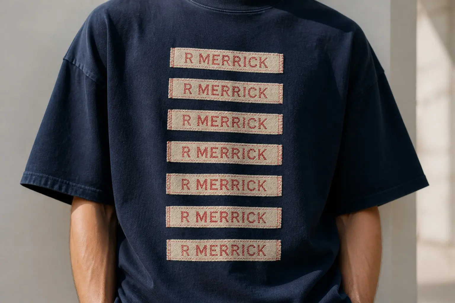 A navy tee printed with seven identical woven name tapes stitched in a single vertical column, one sewn into each of seven edges — collar, cuff, waistband, shoe tongue, bag strap, towel corner, coat hood — every folded rectangle carrying the same two woven capitals, R MERRICK.