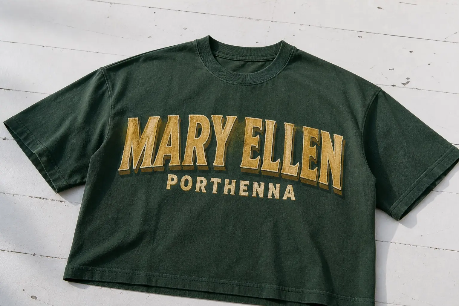 A bottle green shirt printed with the name MARY ELLEN in gold shaded block signwriting with a hard drop shadow, and beneath it in smaller matched lettering the port PORTHENNA, both lines flat with open space at either side.