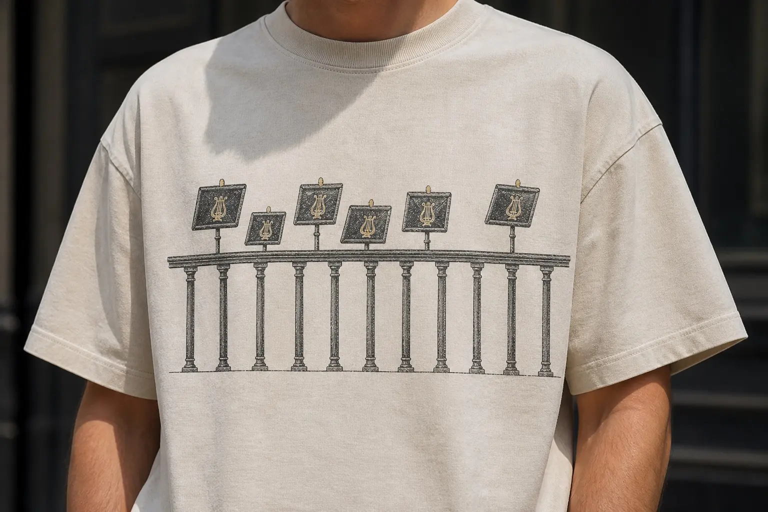 A bone tee printed in charcoal and brass gold with a straight-on section of orchestra pit rail — a plain horizontal top rail on evenly spaced uprights running the shirt's full width — and six music stand tops rising behind it at different heights.