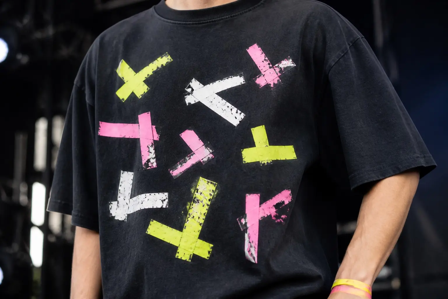 A near-black tee in fluorescent yellow, pink and white printed with eleven spike marks on a dark stage floor seen from directly above — crosses and Ls of gaffer tape at different angles and spacings, several overlapping older marks that have been half peeled up.
